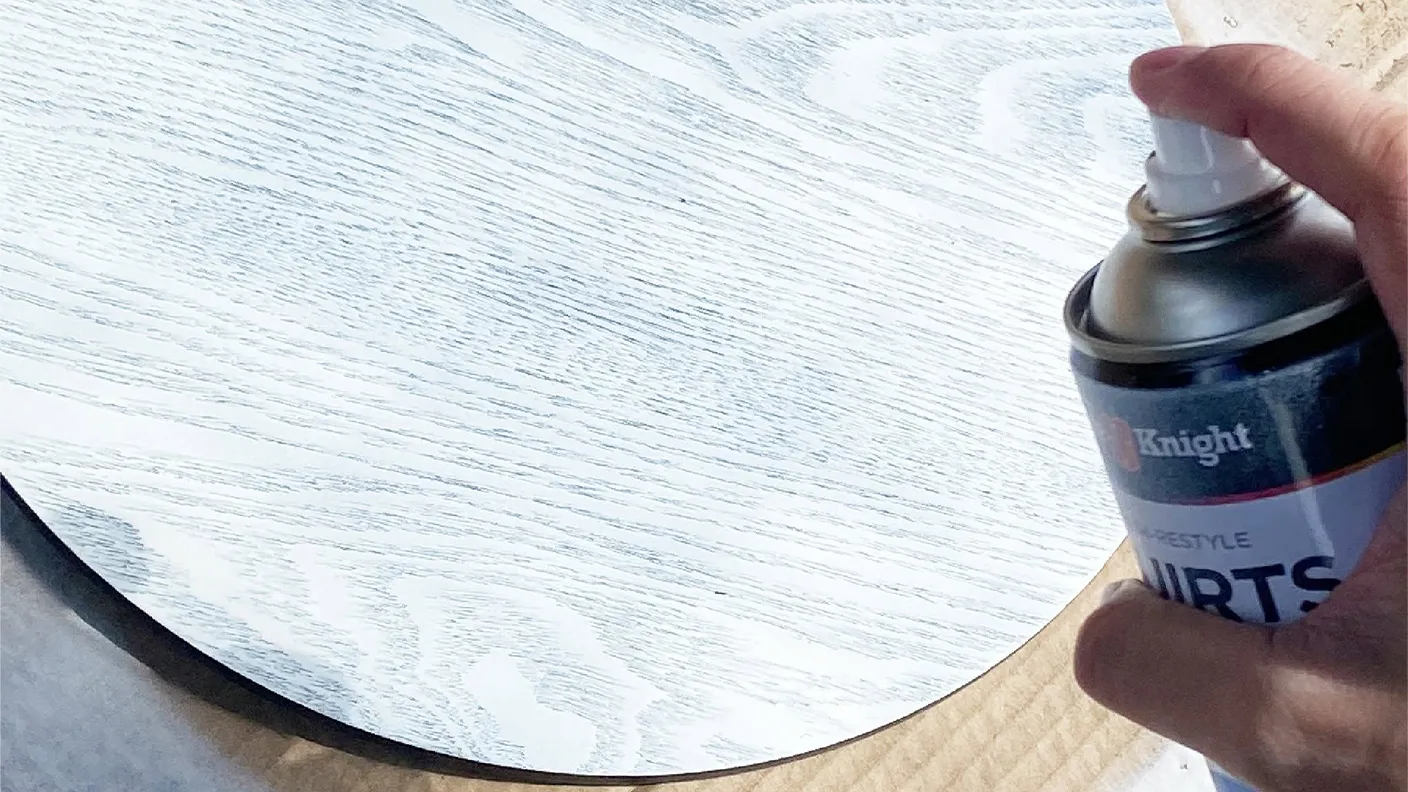A hand holds a spray can, painting a wooden surface with a white finish, highlighting the wood grain texture.