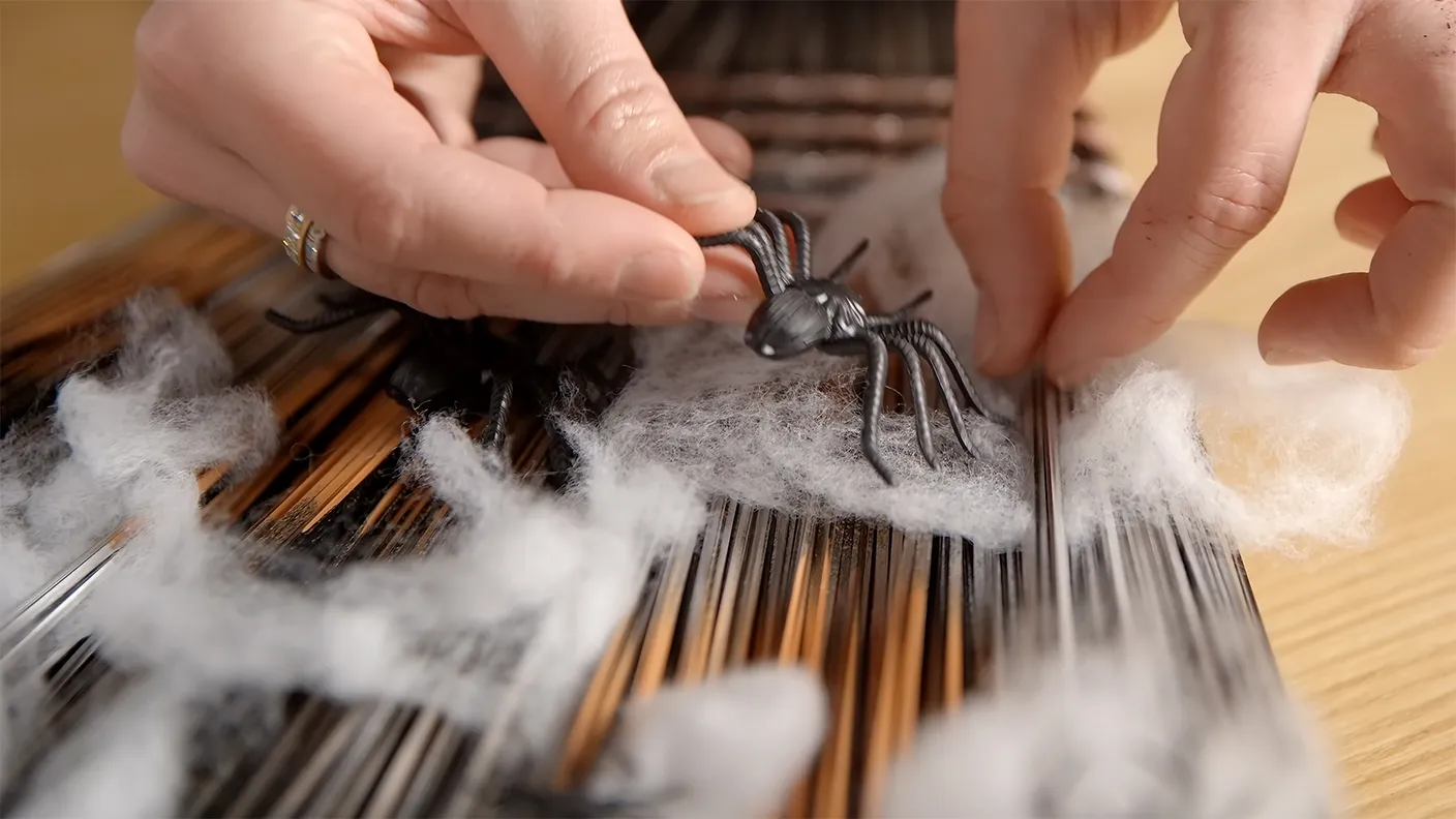 Hands place a plastic spider on a surface with white cotton simulating a web, with wooden sticks in the background.