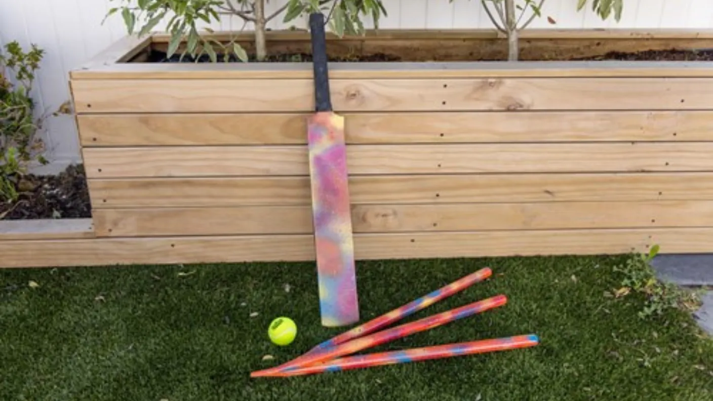 Colorful cricket bat and stumps with a neon yellow ball on grass, leaning against a wooden planter.