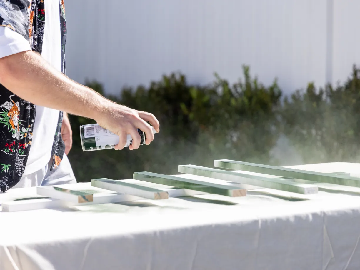 A person spray-paints wooden pieces green on a table outdoors, wearing a floral shirt.