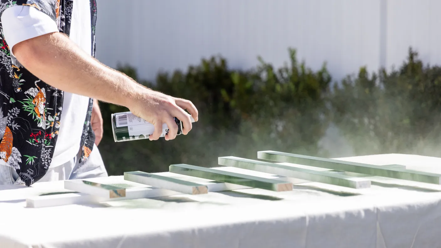 A person spray-paints wooden pieces green on a table outdoors, wearing a floral shirt.