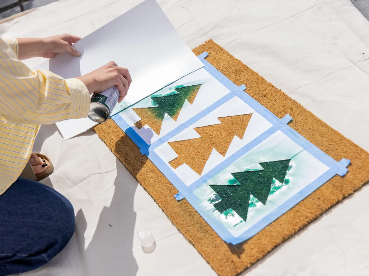 Person spray-painting green Christmas tree stencils on a doormat using blue tape and white paper as guides, outdoors on a sunny day.