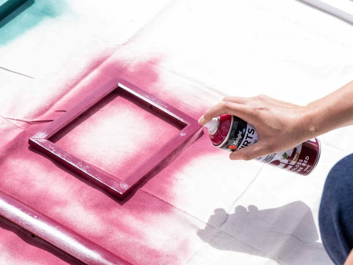 A person spray-painting a picture frame red on a white surface, with green and red paint areas visible.