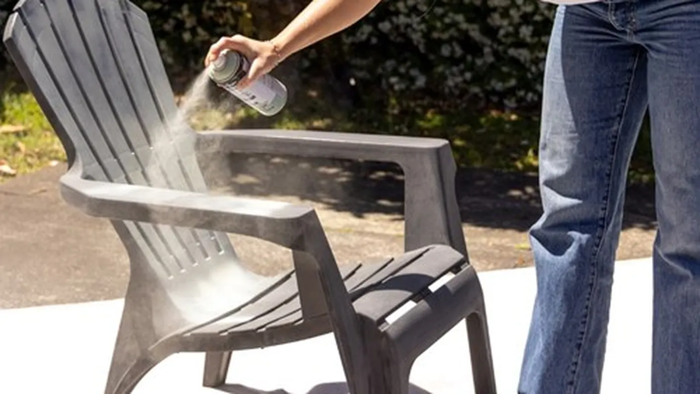 Person spray painting a dark plastic Adirondack chair outdoors, surrounded by sunlight and greenery.