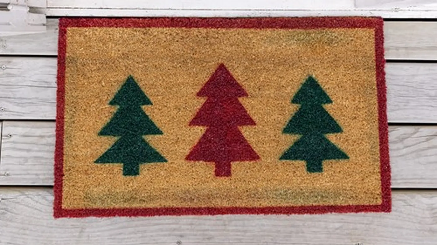 Doormat with two green and one red Christmas tree design on a wooden porch.