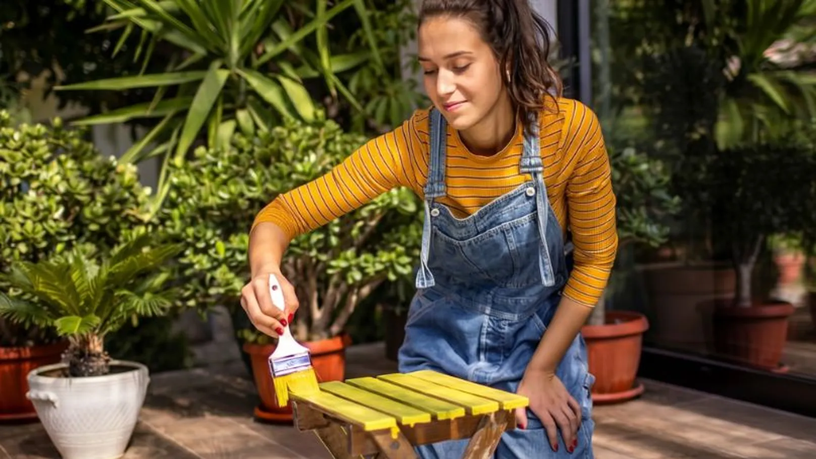 A woman in overalls and a striped shirt paints a small folding table yellow on a sunny patio surrounded by potted plants.