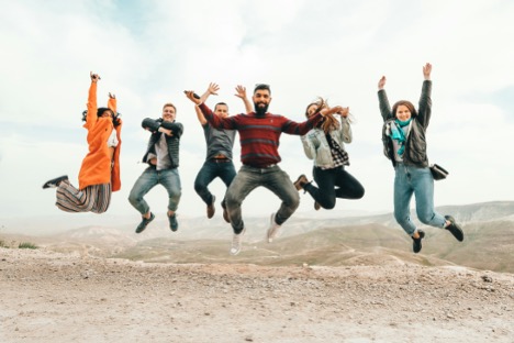Six people jumping in the air on a rocky landscape, expressing joy and excitement under a cloudy sky.