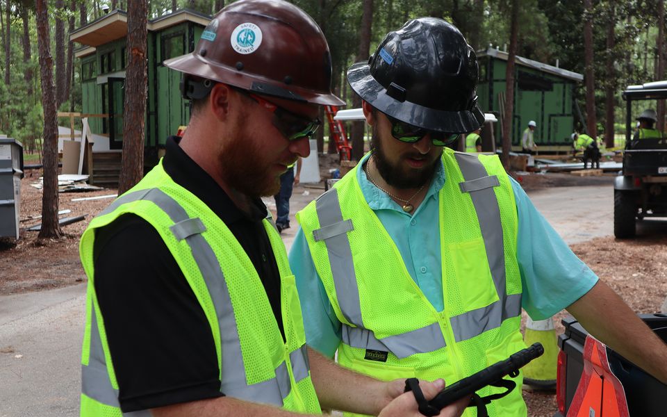 Two workers in high-visibility vests and hard hats discuss plans while holding a tablet at a construction site surrounded by trees and cabins.
