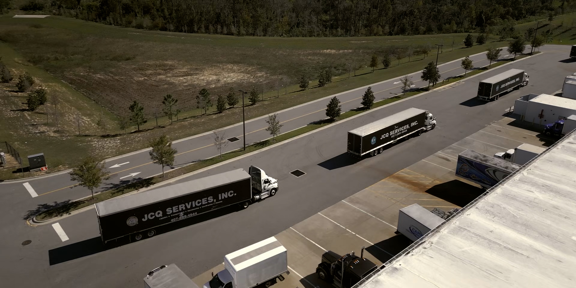Aerial view of JCQ Services, Inc. trucks parked near a warehouse with a road lined by trees and grass fields in the background.