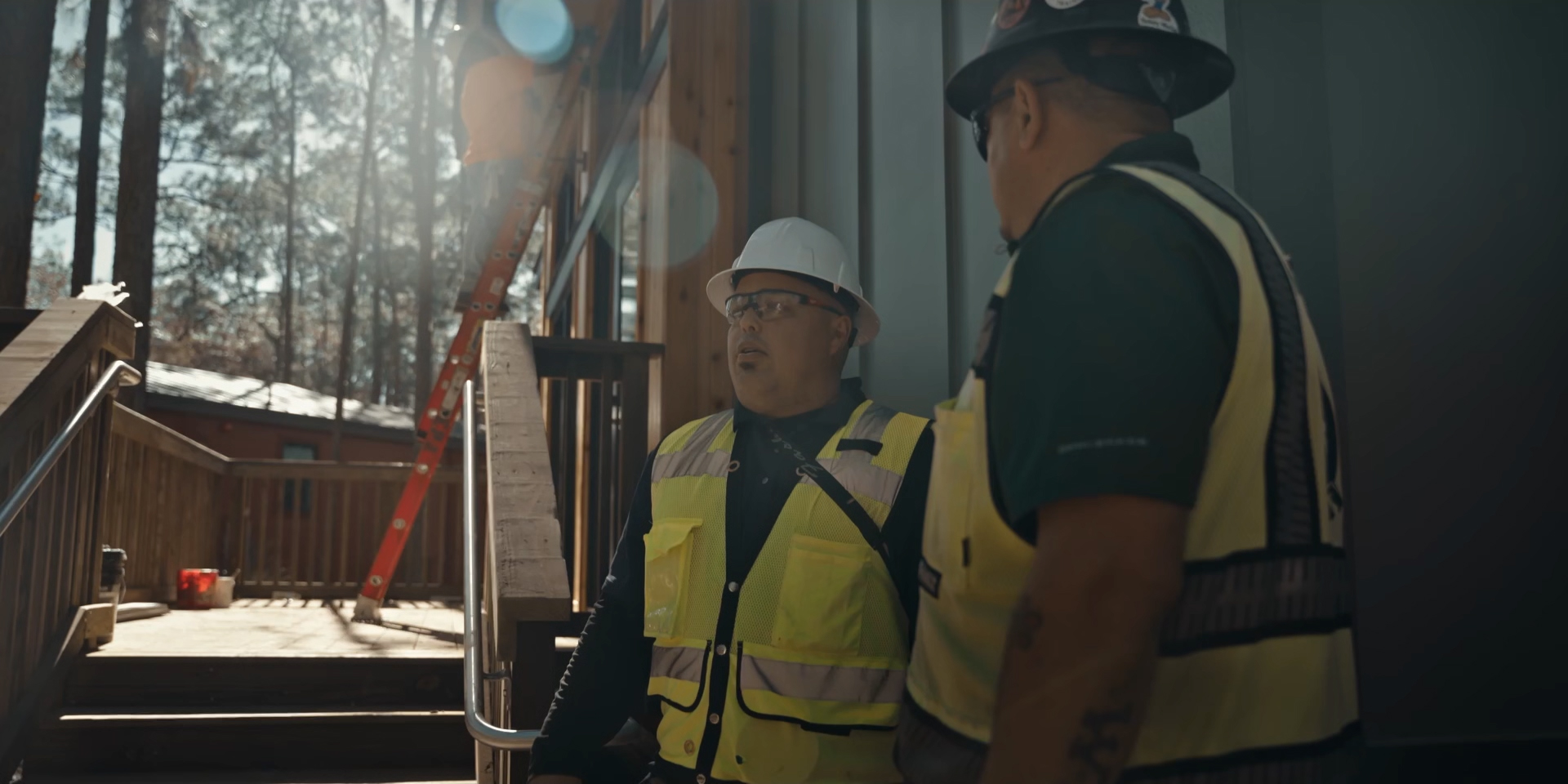 Two construction workers in safety vests and helmets talk outside a building. Sunlight shines through trees, creating lens flare.
