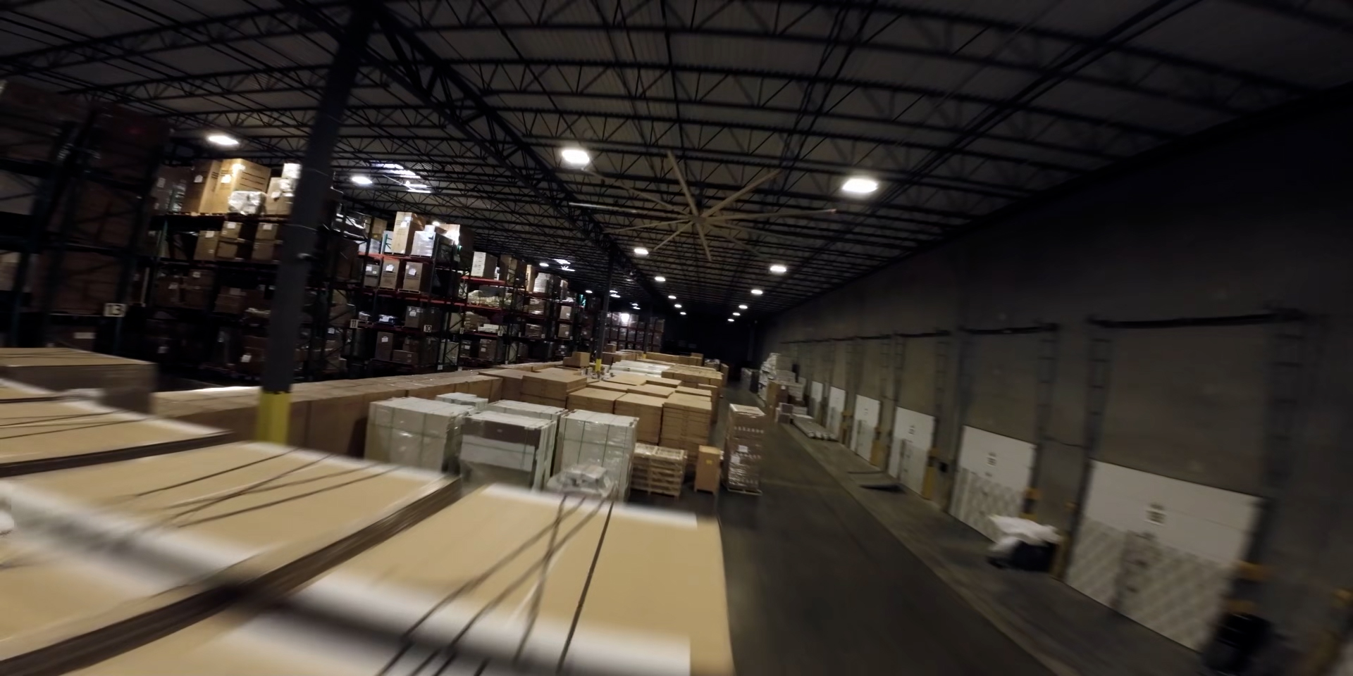 Large, dimly-lit warehouse interior with rows of stacked boxes, pallets, and shelves under a high, metal ceiling.