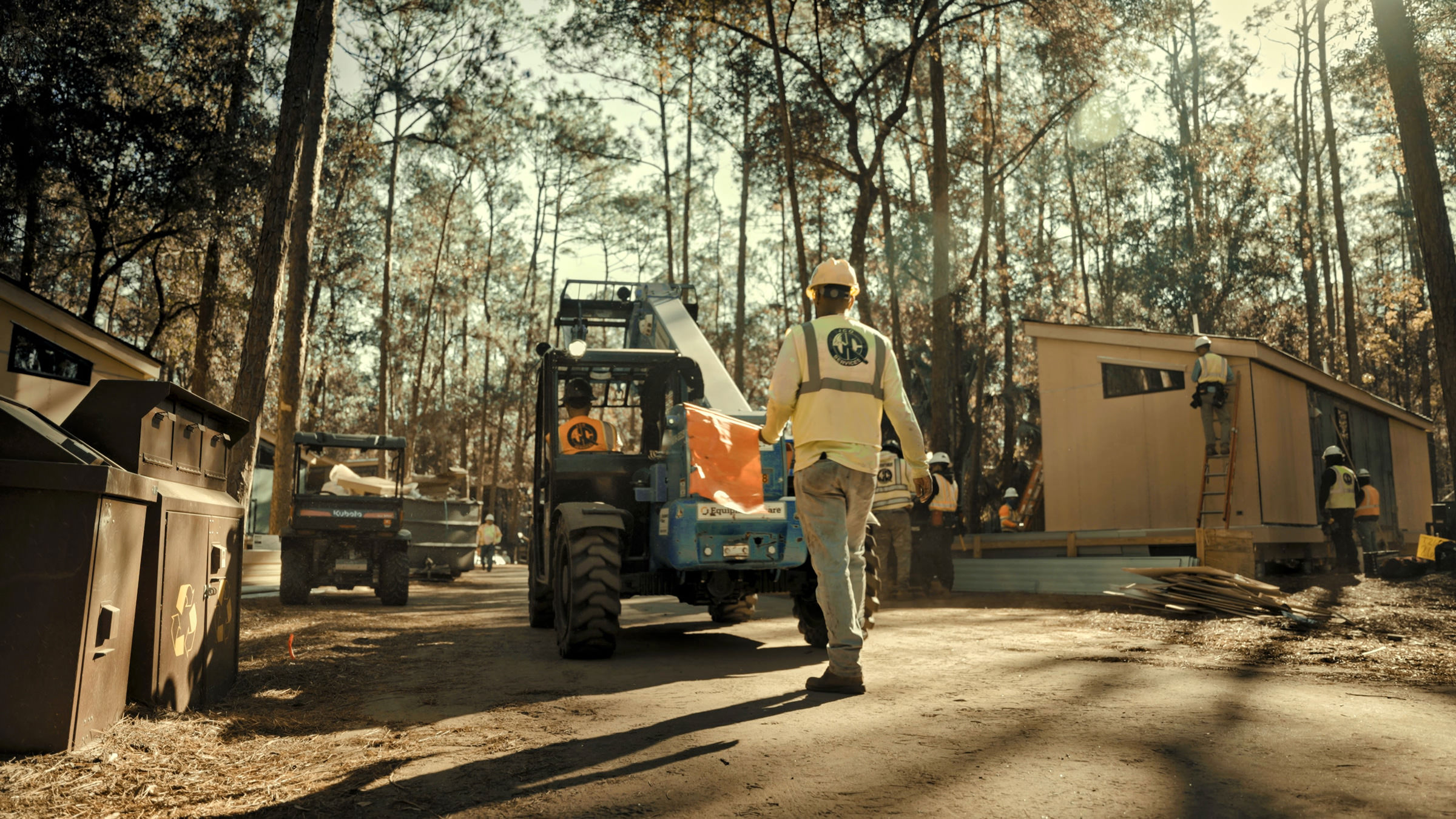 Construction workers on a dirt road operate machinery near a small building, surrounded by trees and sunlight filtering through.