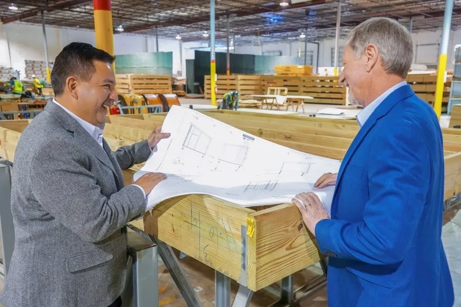 Two men reviewing construction blueprints on wooden beams in a warehouse setting, surrounded by building materials.