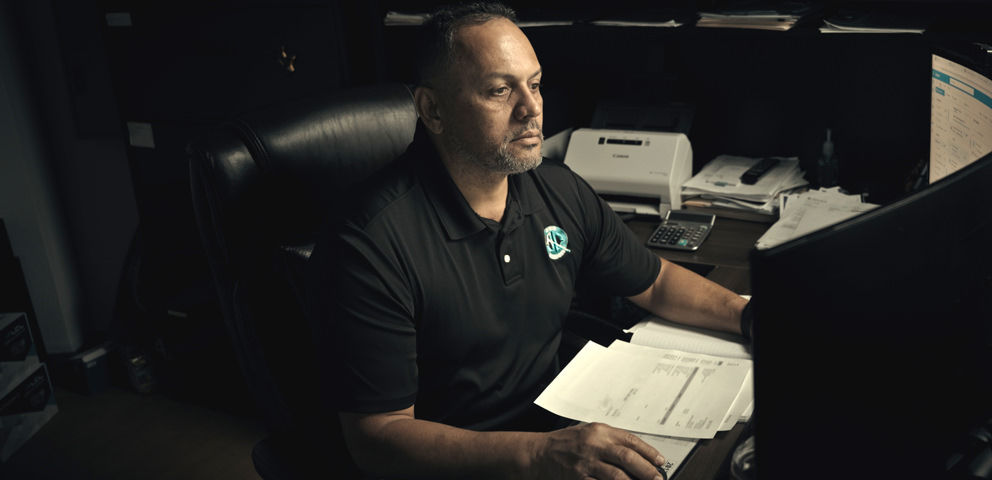 Man in a black polo shirt sits at a desk, focused on a computer monitor, with documents and office supplies around him.