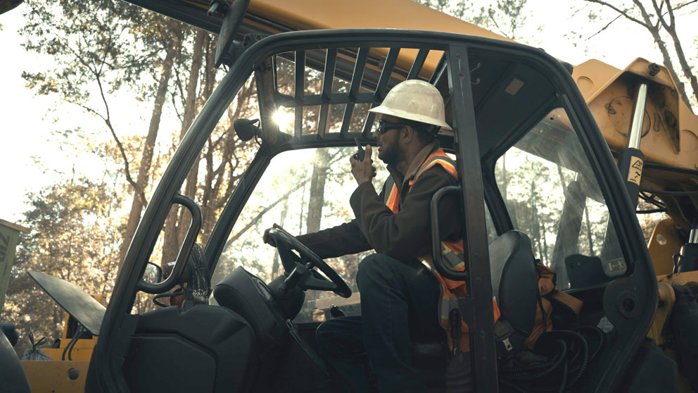 Construction worker in a hard hat and safety vest operates a vehicle, talking on a radio, with trees visible in the background.