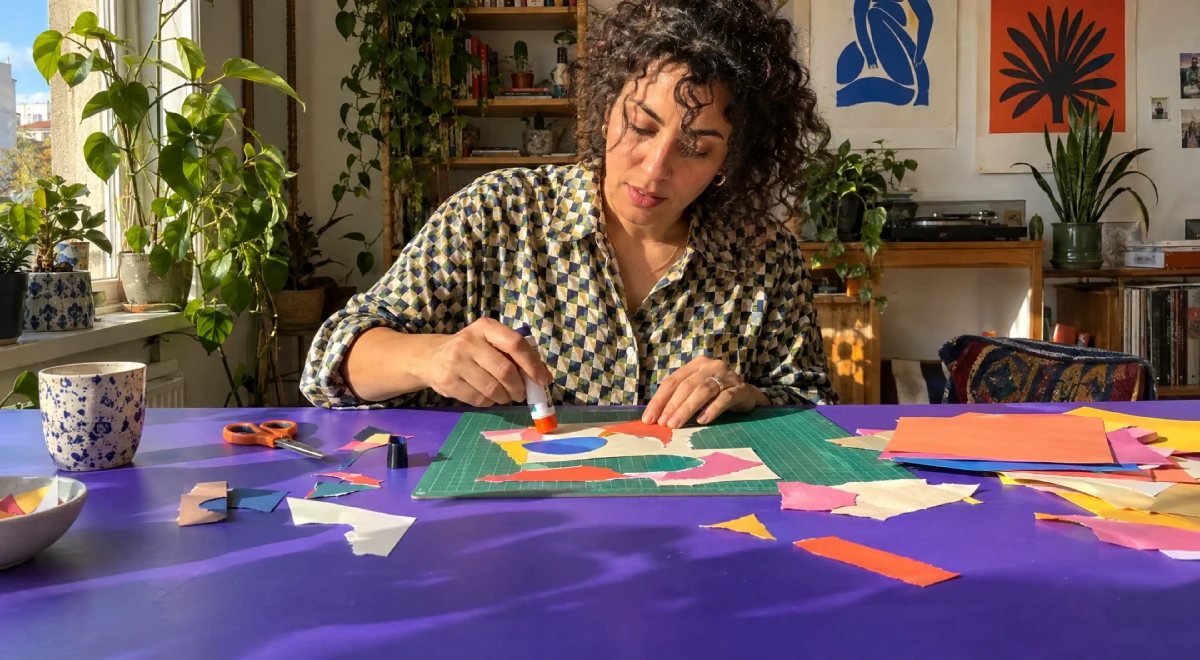 Woman with curly hair enjoying a paper-cutting collage hobby at a purple table with scissors, glue stick and colourful scraps, in a sunny room with plants, books and Matisse-style prints