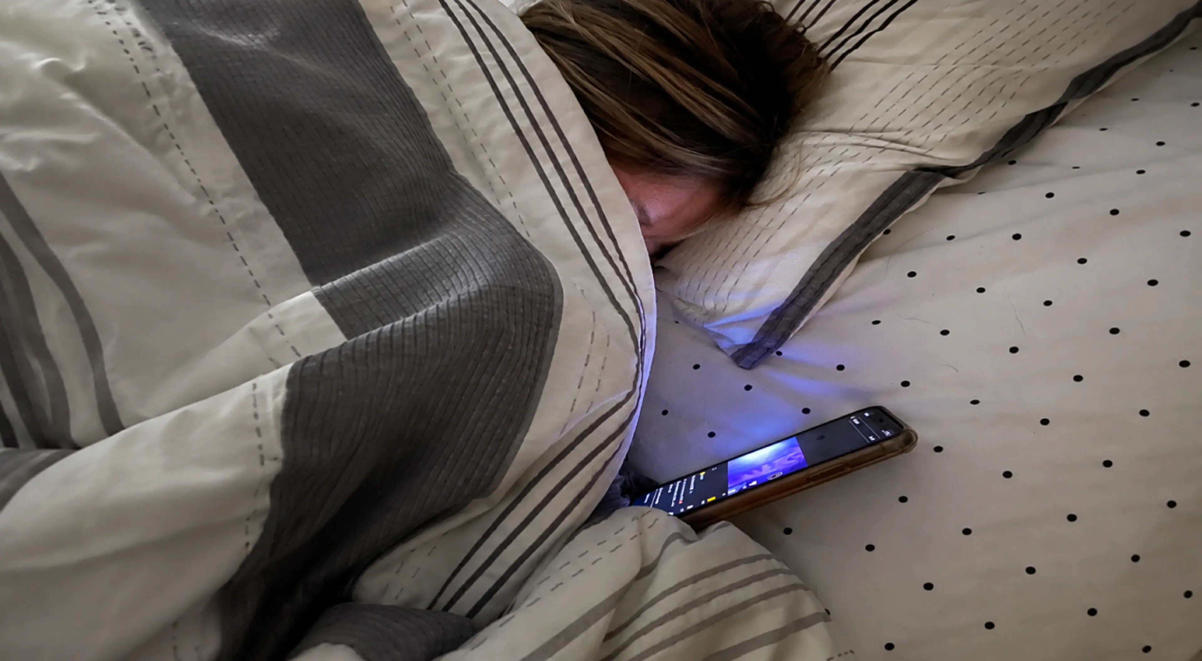 Person lying in bed bored at night, face buried in pillow beside a glowing smartphone on polka-dot sheets under a grays