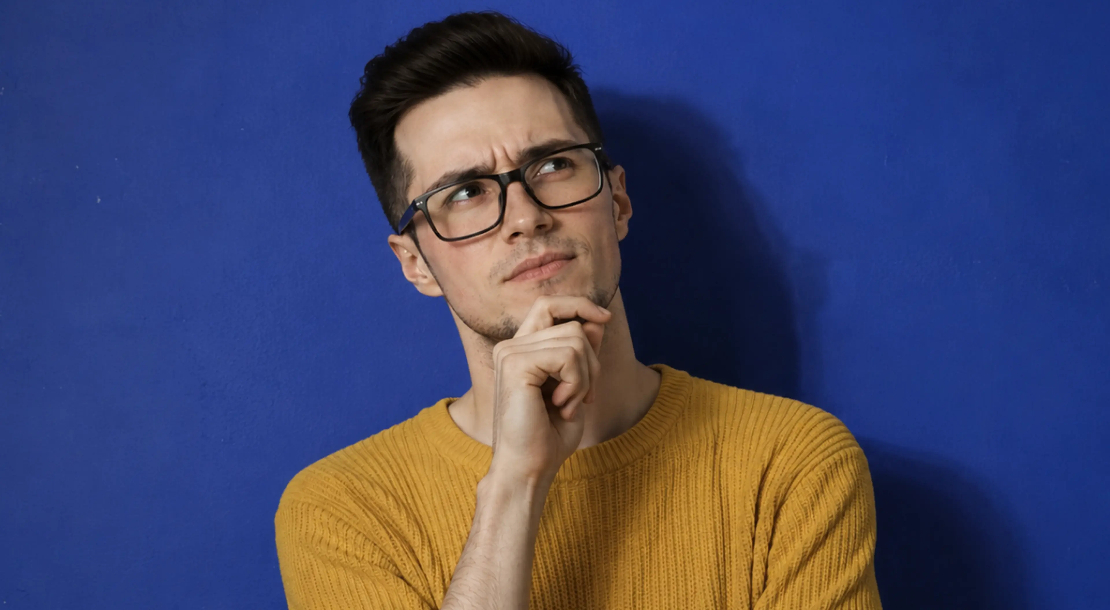 Young man in glasses and a mustard yellow sweater with his hand on his chin, looking up thoughtfully against a blue wall, illustrating critical thinking