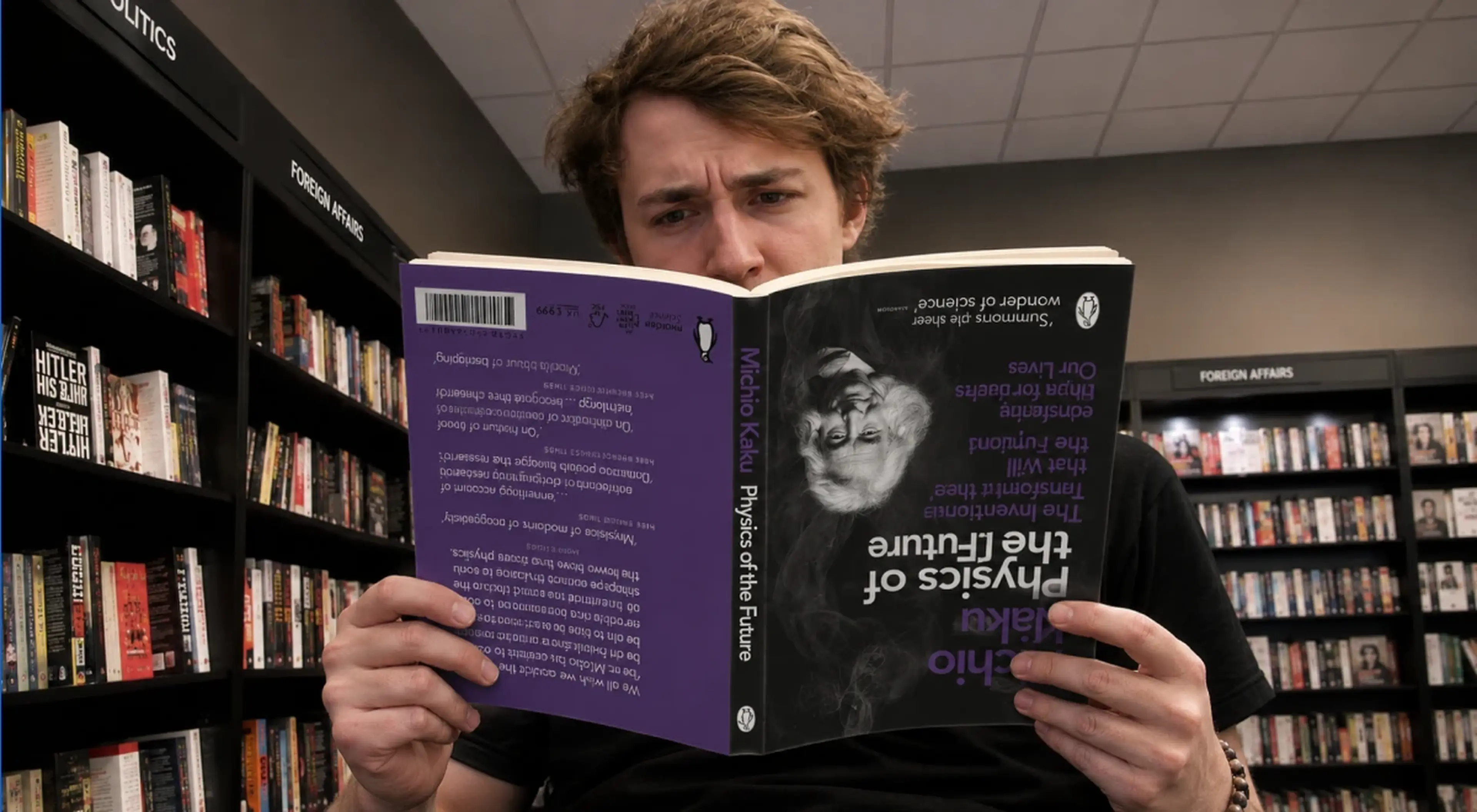Young man reading Physics of the Future by Michio Kaku in a bookstore with shelves of books in the background