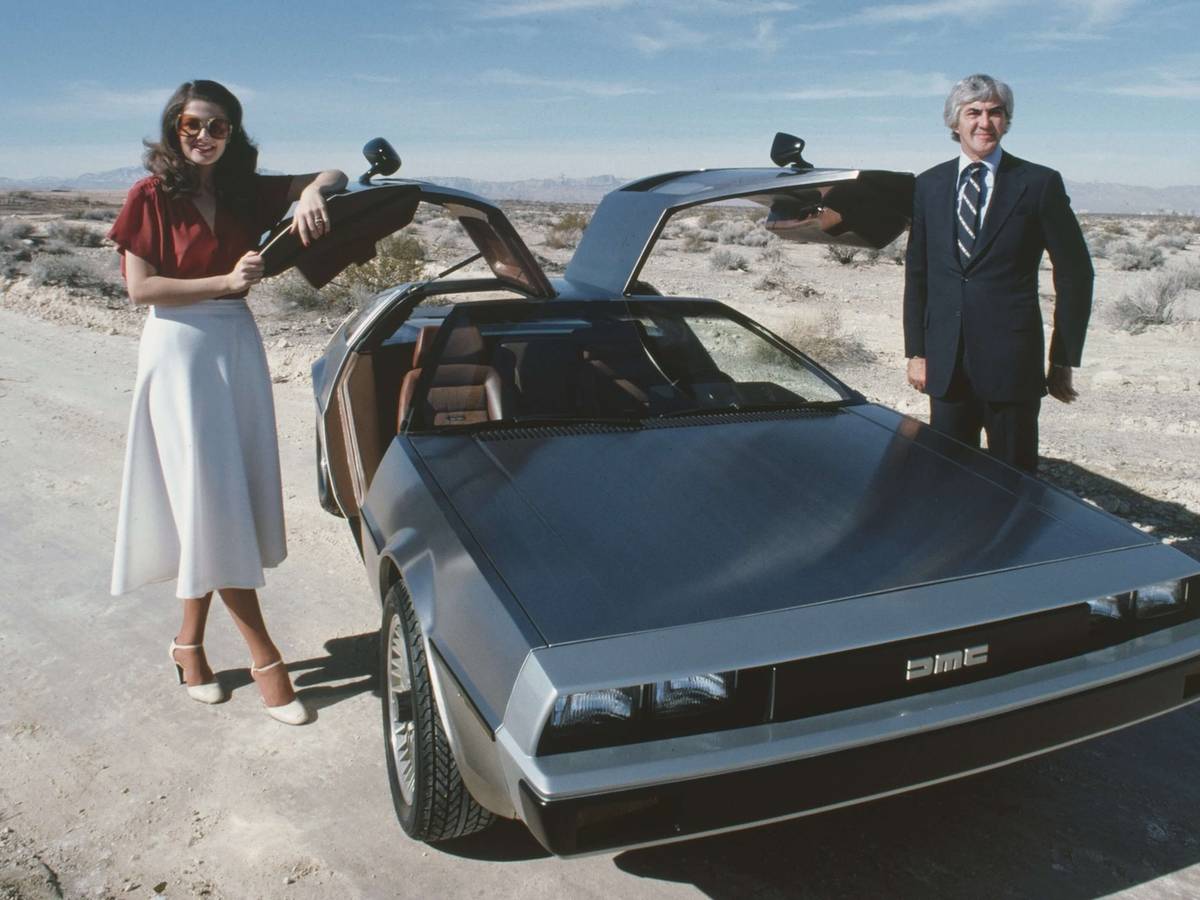 A man and woman stand beside a DeLorean car with open gull-wing doors on a desert road.