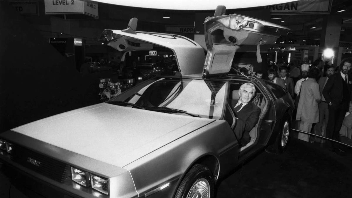 A man in a suit sits in a DeLorean car with gull-wing doors open, surrounded by people at a car show. The setting is in black and white.