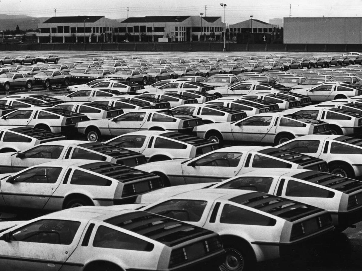 A large parking lot filled with rows of identical DeLorean cars with open doors, set against a backdrop of industrial buildings. Black and white photo.