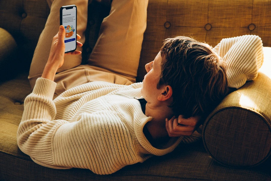 Frau liegt auf dem Sofa und schaut auf ihr Handy. Auf dem Bildschirm sieht sie den Startbildschirm von hiToco für den Trainingsplan.