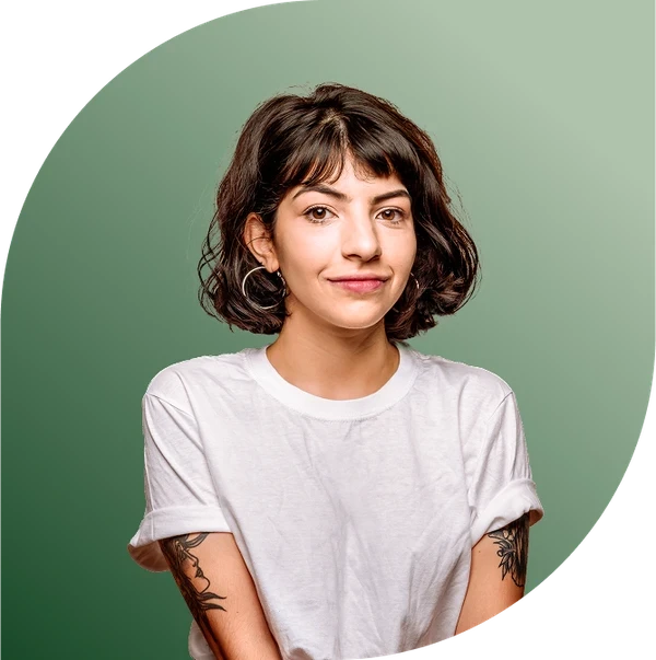 Eine Frau mit kurzen braunen Haaren, die ein weißes T-Shirt trägt, steht vor einem grünen Hintergrund und lächelt sanft. Sie hat Tätowierungen auf ihren Armen.