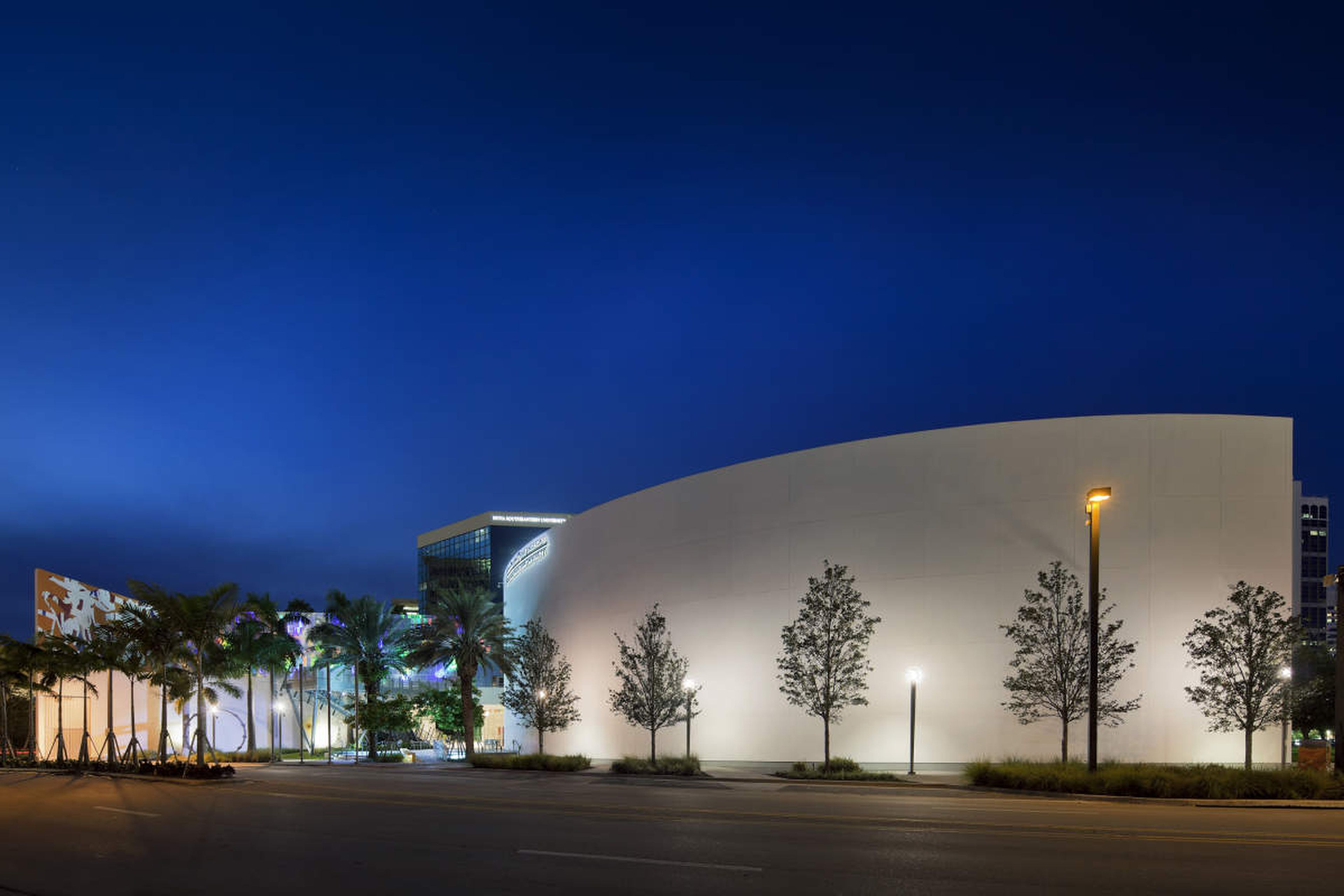 Modern building with curved white facade illuminated at night, surrounded by trees and colorful accent lighting against a deep blue sky.
