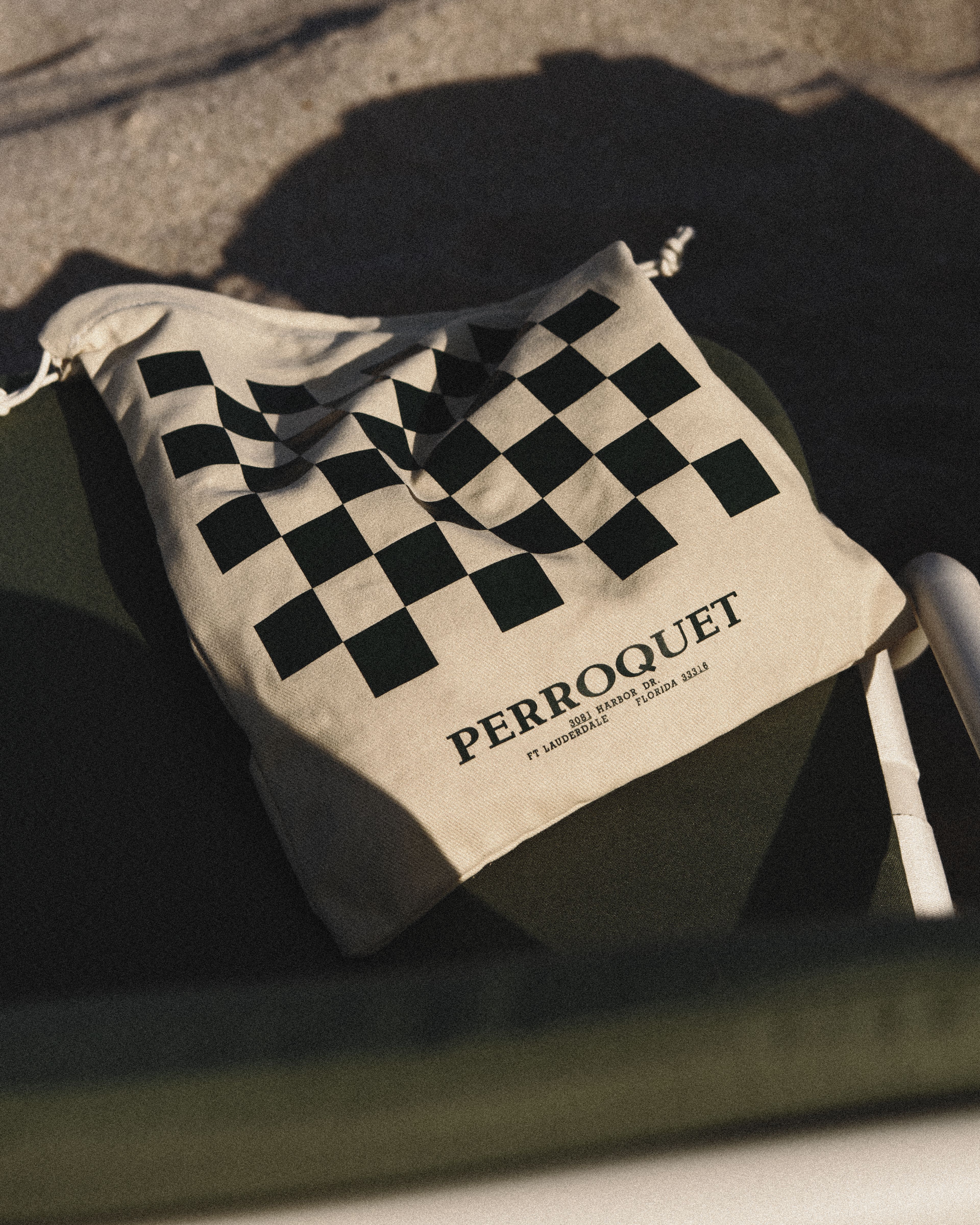 A black-and-white checkered drawstring pouch printed with the Perroquet logo and Fort Lauderdale address, resting on a chair in golden-hour light.