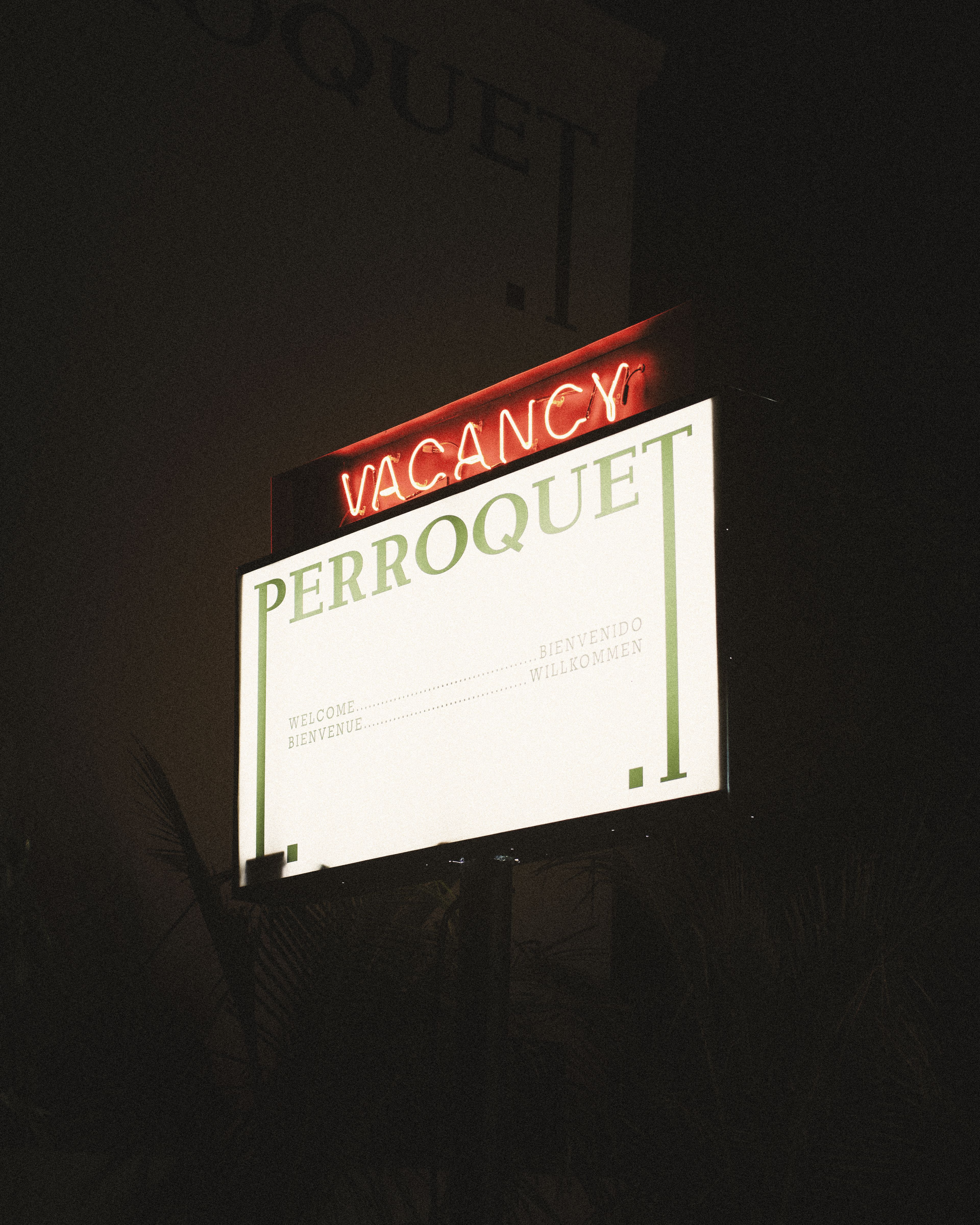 Nighttime shot of the illuminated Perroquet sign with a red "Vacancy" neon light above the hotel's welcome message in multiple languages.