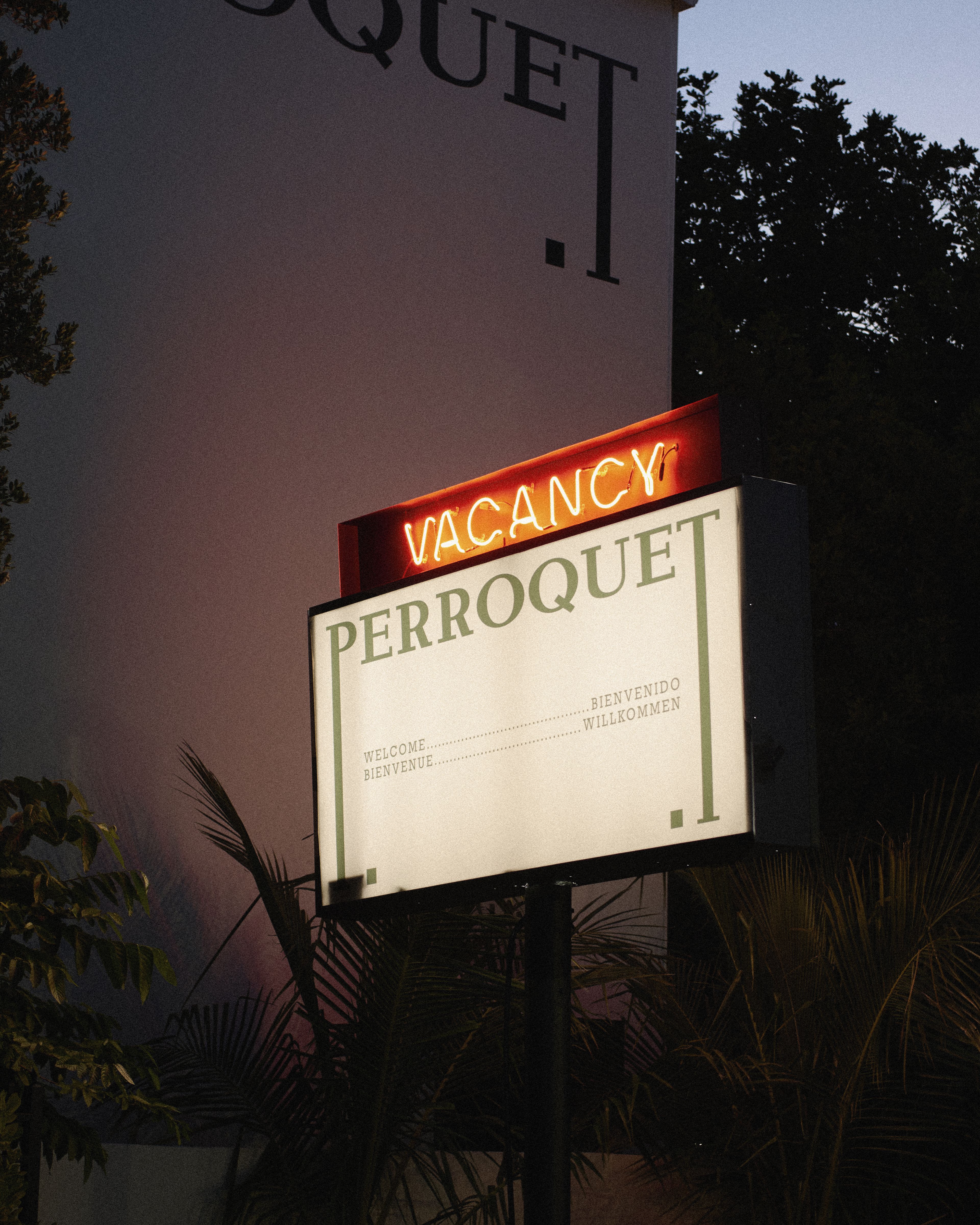 Dusk view of the Perroquet sign glowing beneath the hotel's painted logo on the building facade, framed by trees and palms.