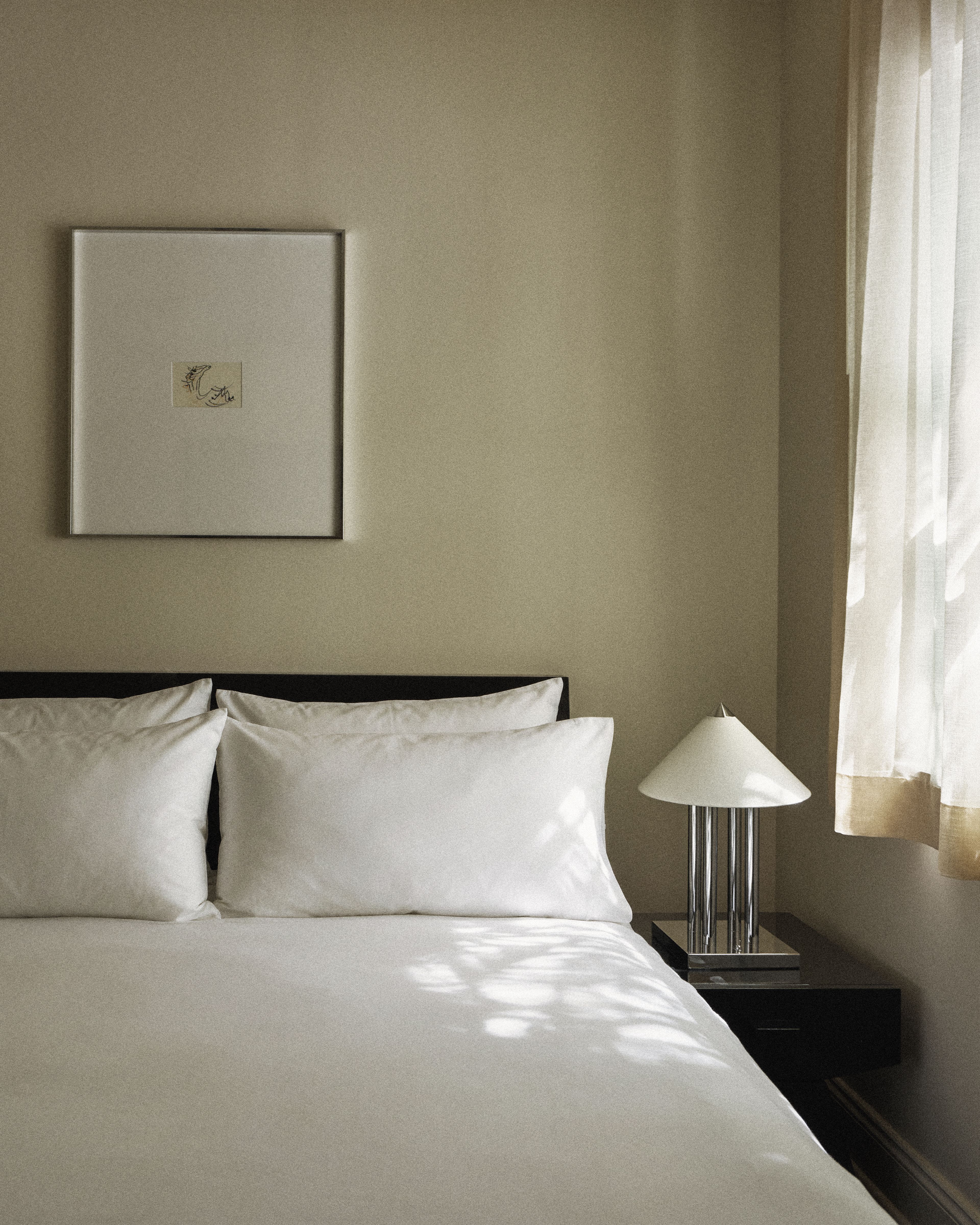 Close-up of bed, modern bedside table, and soft natural light filtering through sheer curtains.