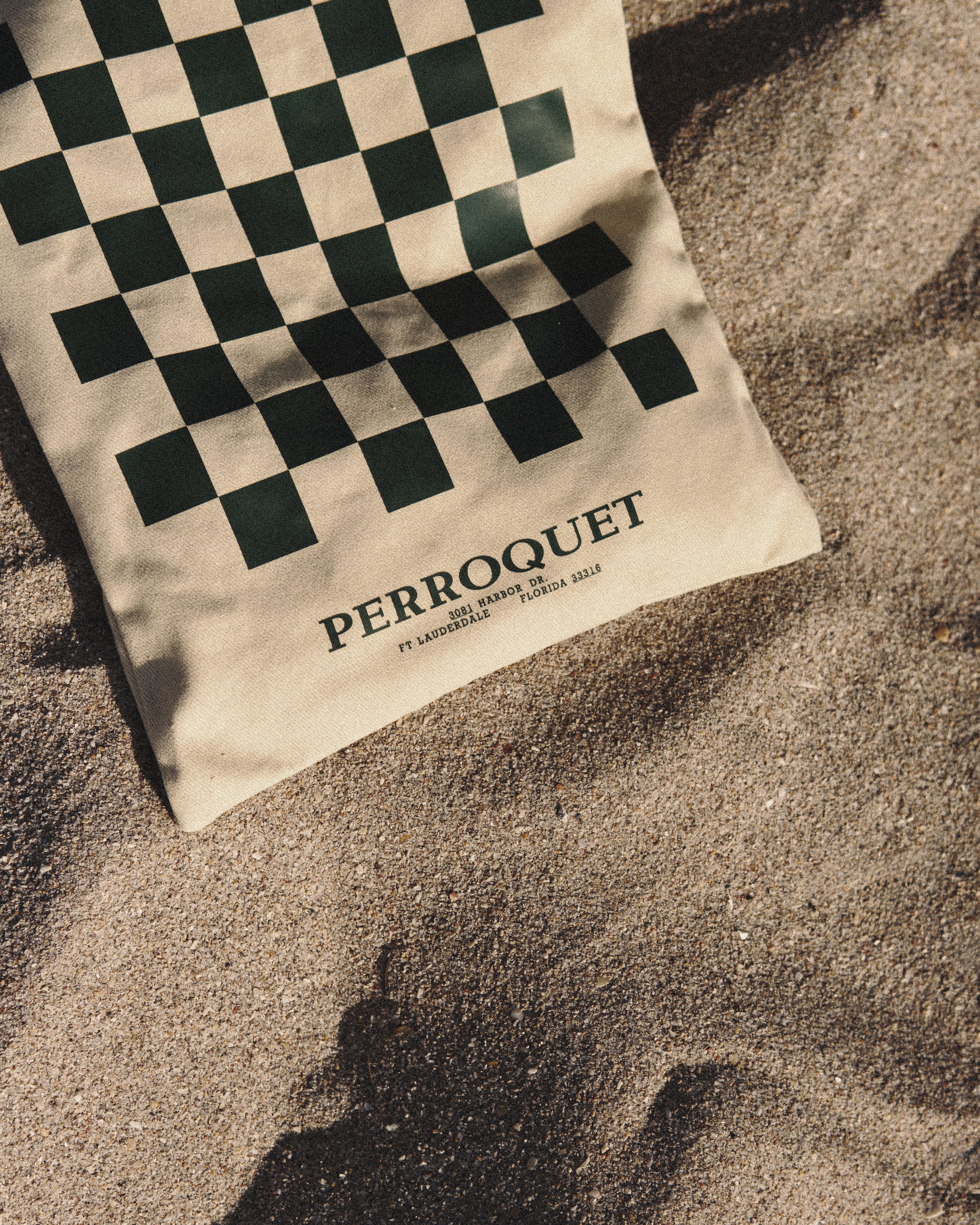 Close-up of a green-and-cream checkered Perroquet tote bag lying on sand, showing the "Perroquet, Ft Lauderdale, Florida 33316" address print.