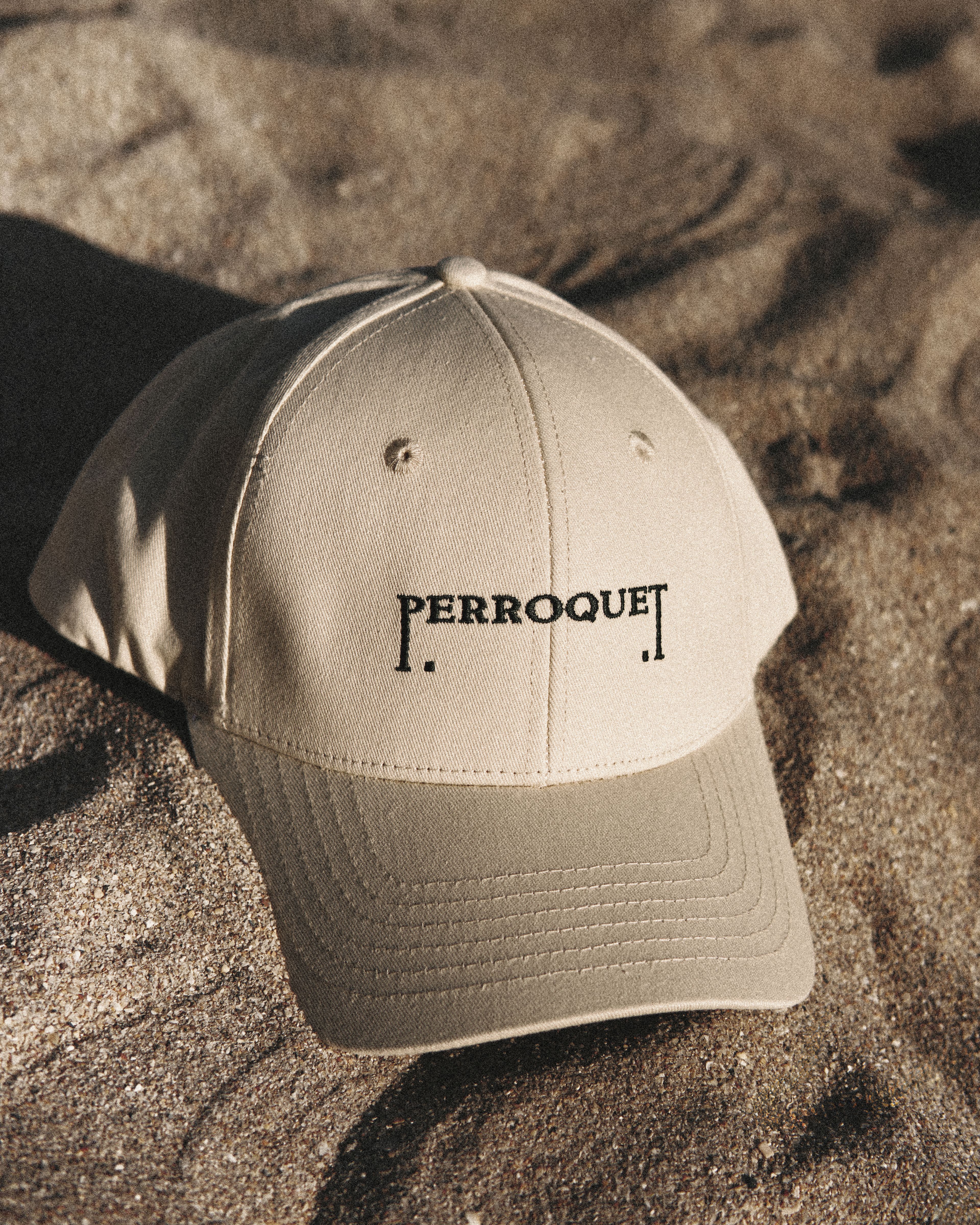 A tan baseball cap embroidered with the Perroquet logo, resting on sand in warm sunlight.