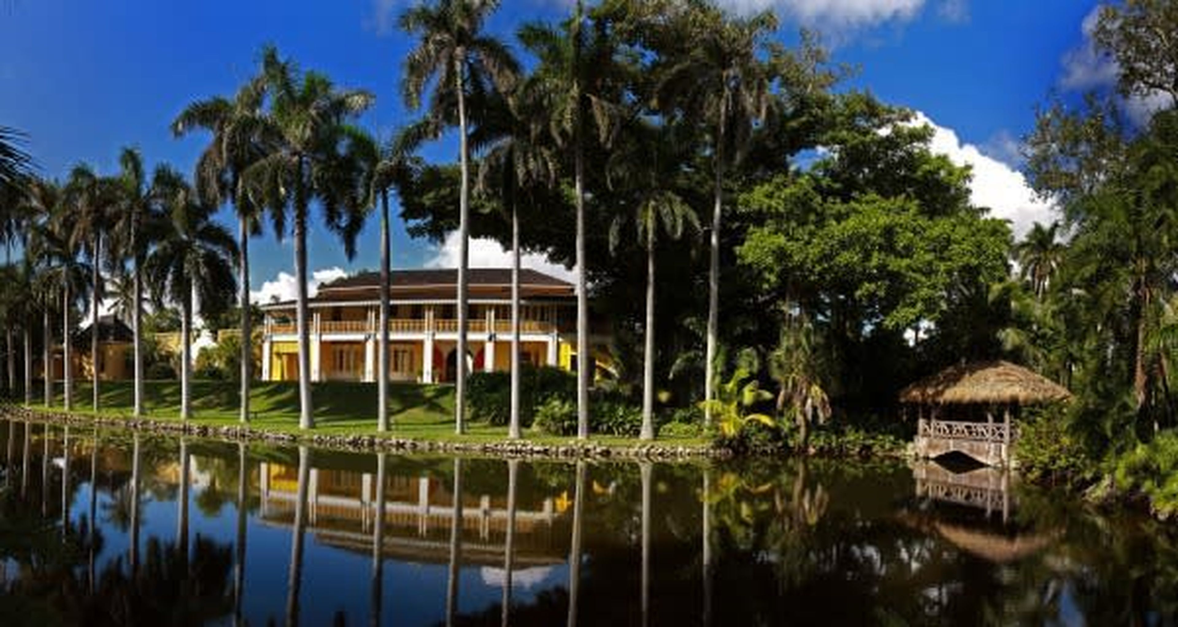 Elegant yellow building with tall columns, surrounded by palm trees, reflected in a tranquil pond with a rustic gazebo on the right.