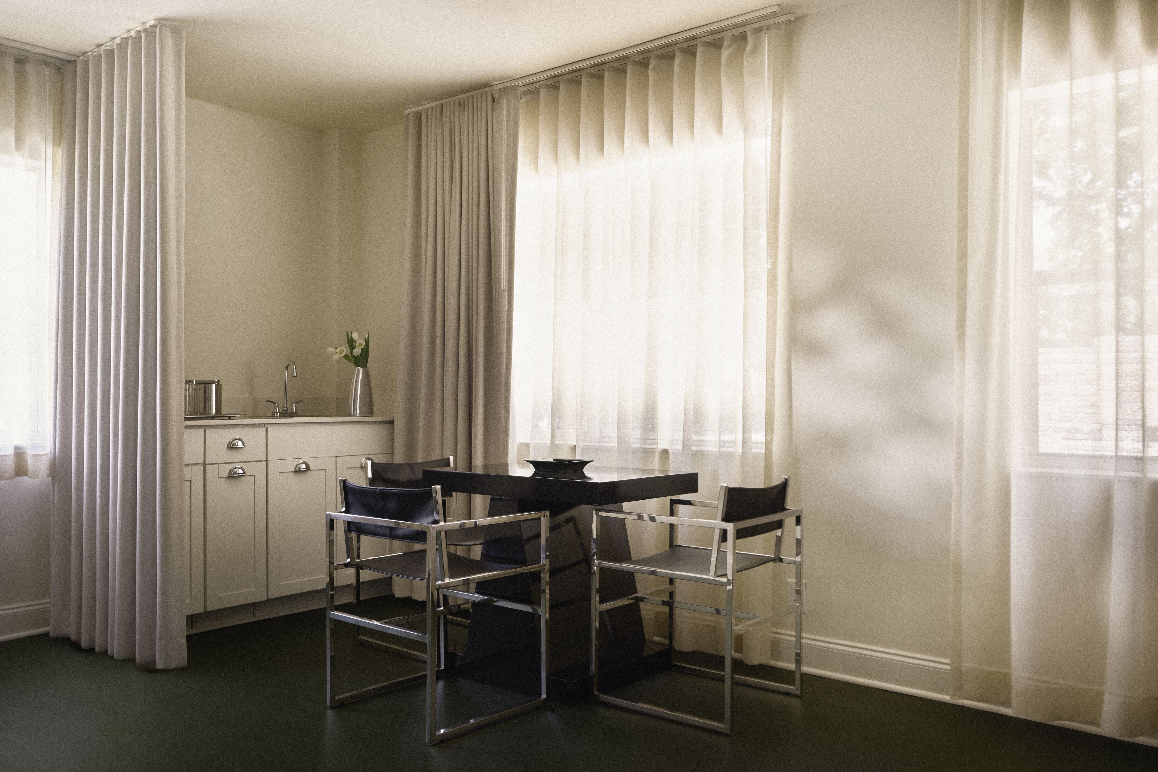 Sitting area with dining table, kitchenette, and floor-to-ceiling sheer curtains in a Perroquet Hotel guest room.