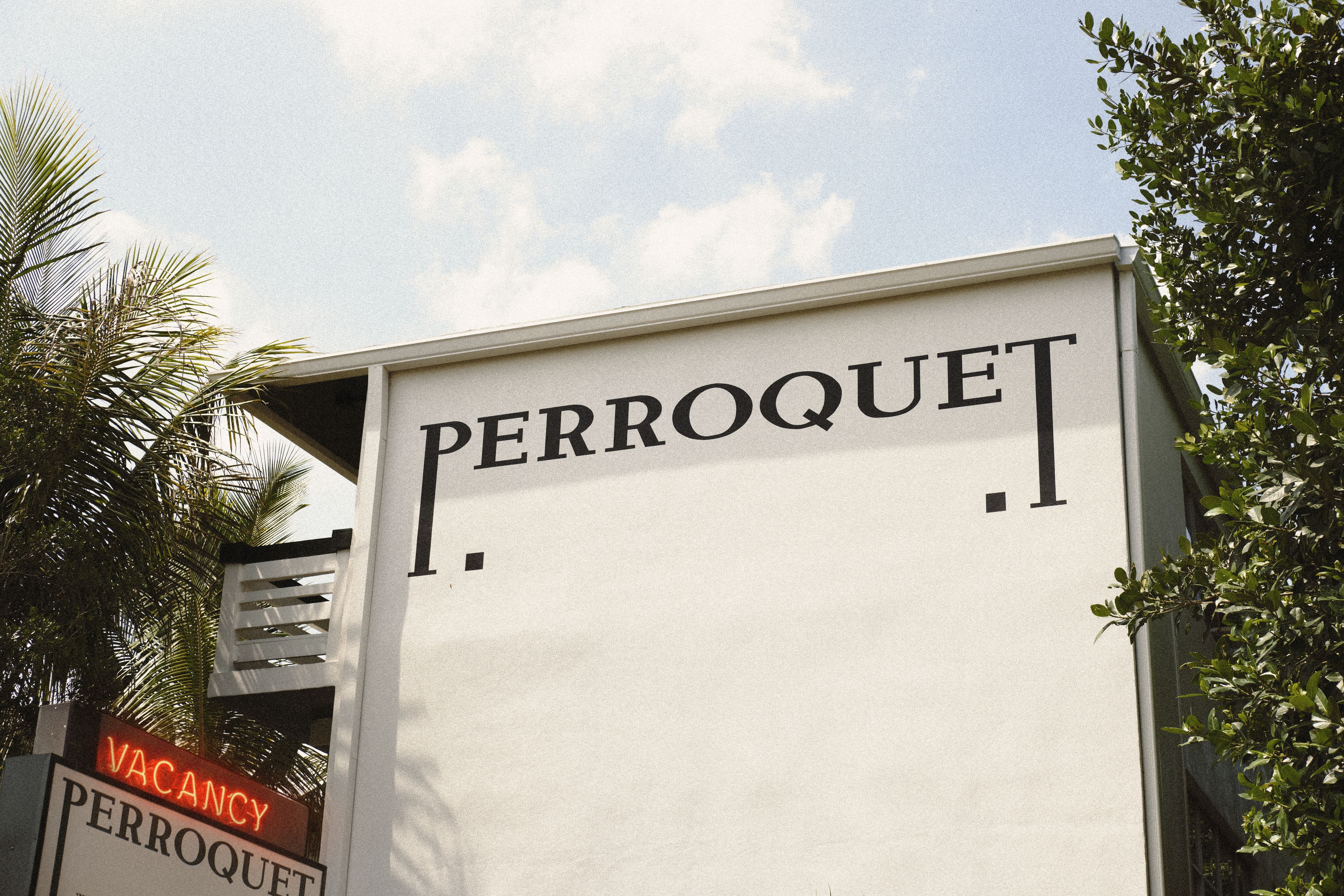 Entrance to Perroquet Hotel in Fort Lauderdale with its signature black logo on a white façade, surrounded by palm trees and lush tropical landscaping.