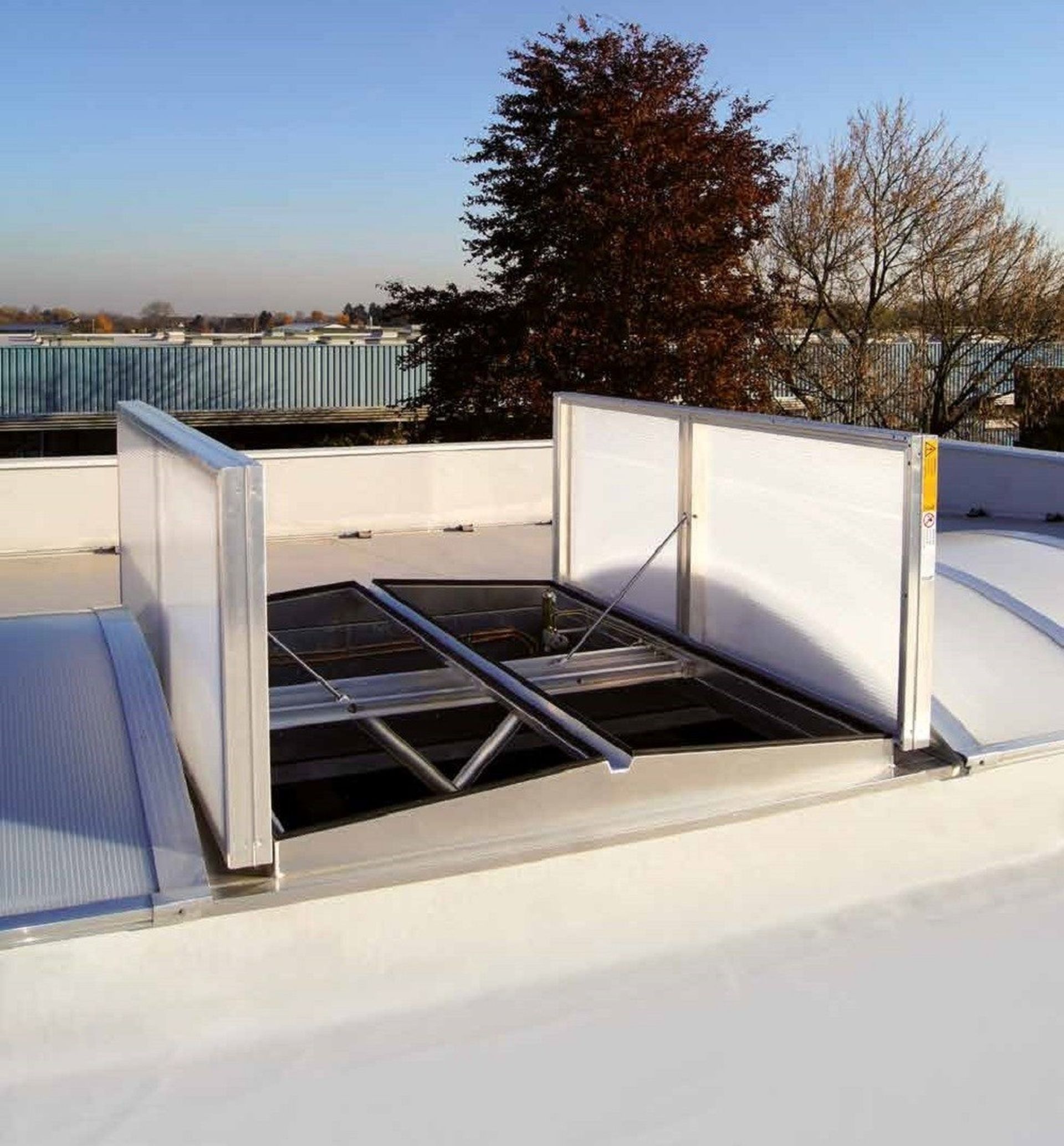 Open roof ventilation hatch on a flat rooftop, surrounded by trees and industrial buildings in the background under a clear sky.