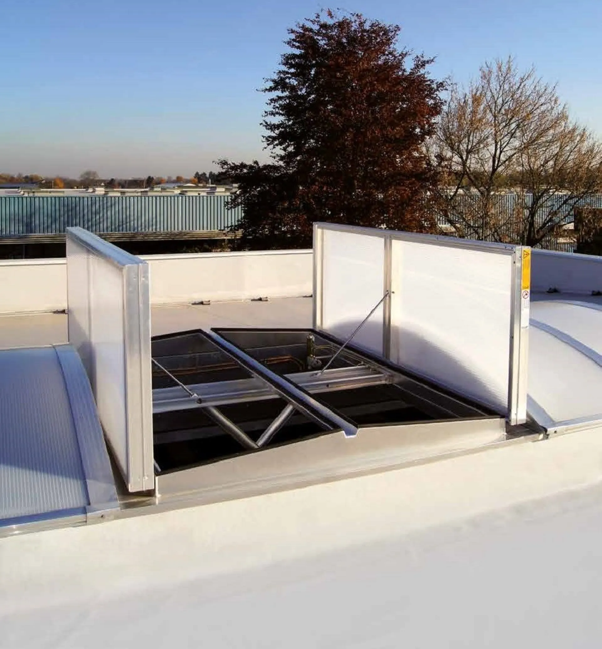 Open roof ventilation hatch on a flat rooftop, surrounded by trees and industrial buildings in the background under a clear sky.