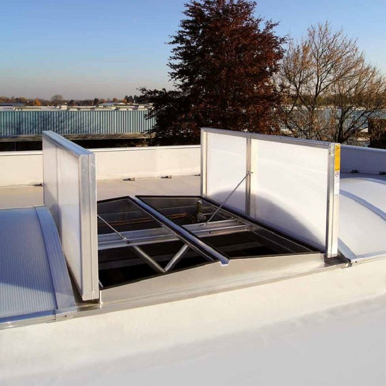 Open roof ventilation hatch on a flat rooftop, surrounded by trees and industrial buildings in the background under a clear sky.