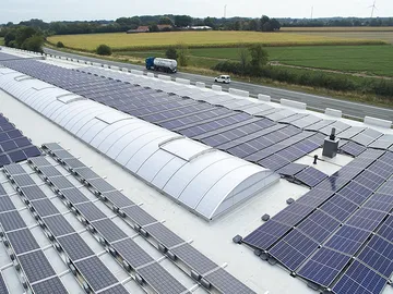 Rooftop solar panels and Continuous rooflight LB basic on a large building near a country road, surrounded by fields and greenery under a clear sky.