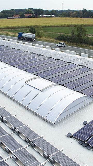 Rooftop solar panels and Continuous rooflight LB basic on a large building near a country road, surrounded by fields and greenery under a clear sky.