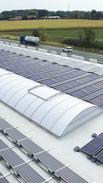 Rooftop solar panels and Continuous rooflight LB basic on a large building near a country road, surrounded by fields and greenery under a clear sky.