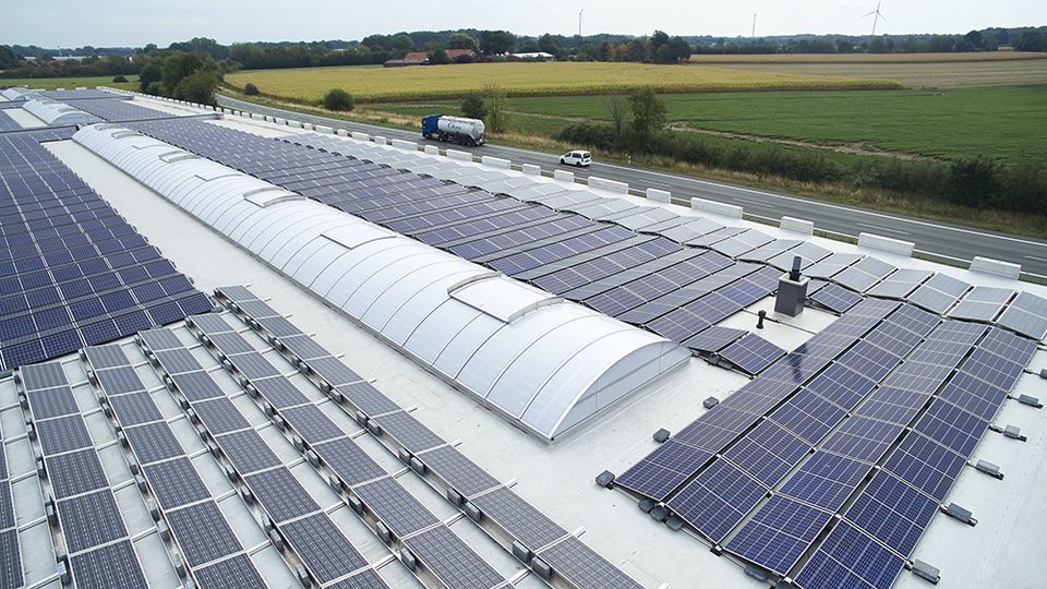 Rooftop solar panels and Continuous rooflight LB basic on a large building near a country road, surrounded by fields and greenery under a clear sky.