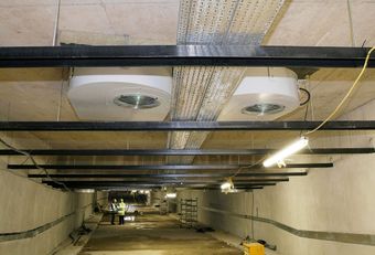 An underground tunnel with installed ventilation systems on the ceiling, metal beams, and two workers wearing safety vests and helmets.