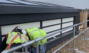 Two construction workers in safety gear are inspecting the base of a smoke control system, surrounded by pebbles and safety rails.