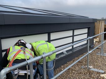 Two construction workers in safety gear are inspecting the base of a smoke control system, surrounded by pebbles and safety rails.