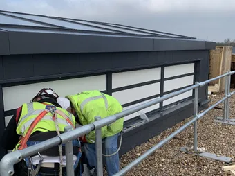 Two construction workers in safety gear are inspecting the base of a smoke control system, surrounded by pebbles and safety rails.