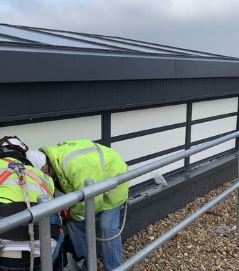 Two construction workers in safety gear are inspecting the base of a smoke control system, surrounded by pebbles and safety rails.